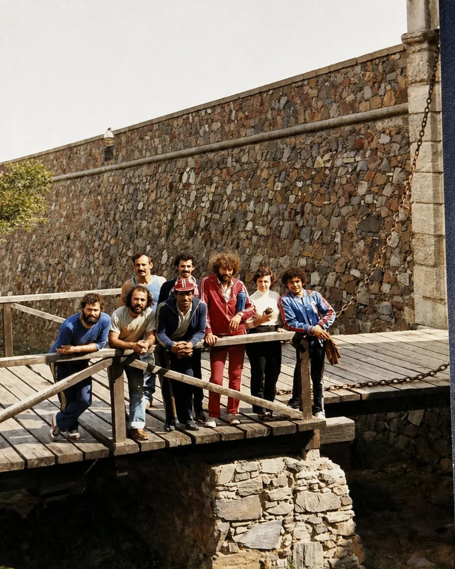 Ocho personas posando sobre el puente de madera de entrada a una fortaleza. Detrás se ve la muralla de piedra y la cadena del puente levadizo; uno de ellos se apoya en un bastón.