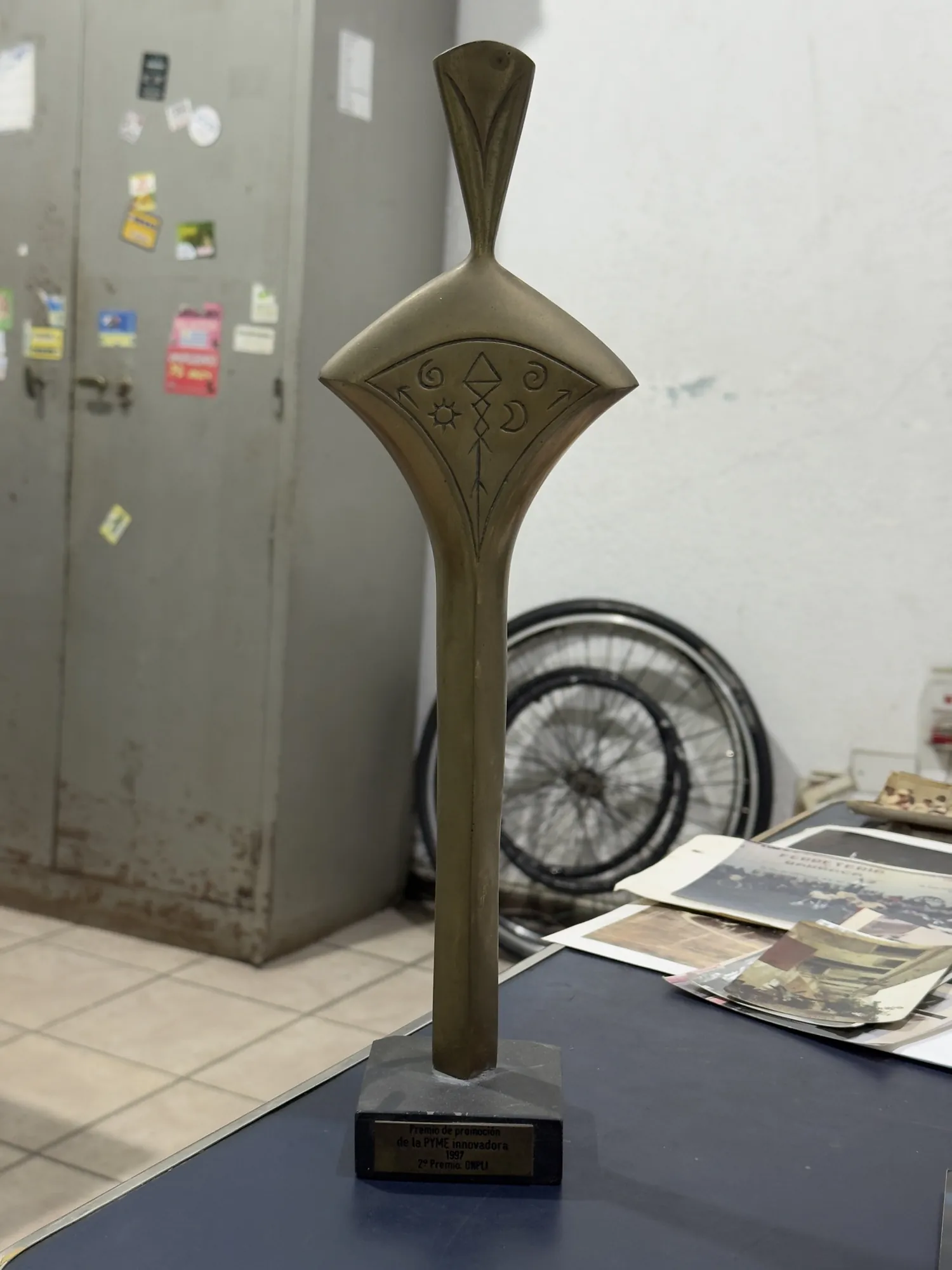 Trofeo de bronce con una figura estilizada y símbolos grabados, apoyado sobre una base de piedra con una placa negra en el frente.