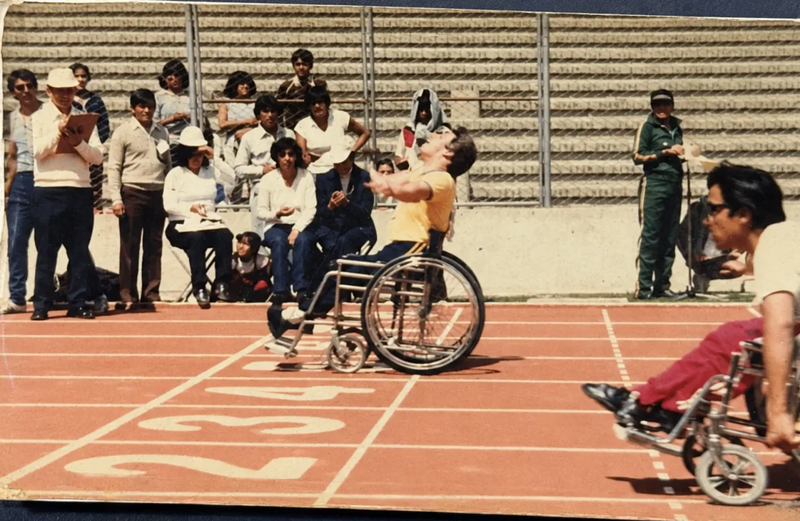 Competencia de atletismo en pista: un atleta en silla de ruedas ejecuta un lanzamiento mientras otro compite en un carril vecino, con jueces y público siguiendo la prueba.
